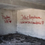 Interior of an abandoned room with white walls covered in red graffiti and hearts; messages include 'I think that life is beautiful' amid rubble on the floor.