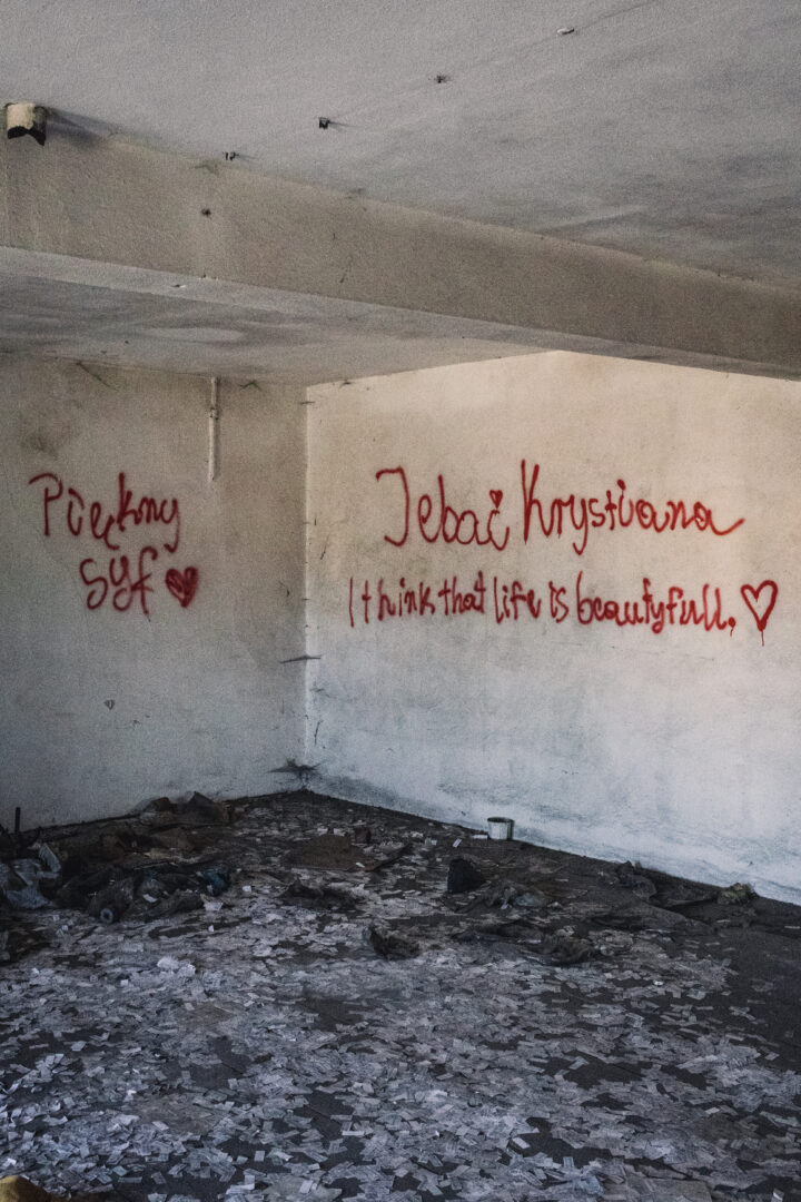 Interior of an abandoned room with white walls covered in red graffiti and hearts; messages include 'I think that life is beautiful' amid rubble on the floor.