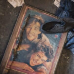 A torn poster of a smiling couple lying on a dusty floor, with a black boot resting on the frame and scattered papers nearby.