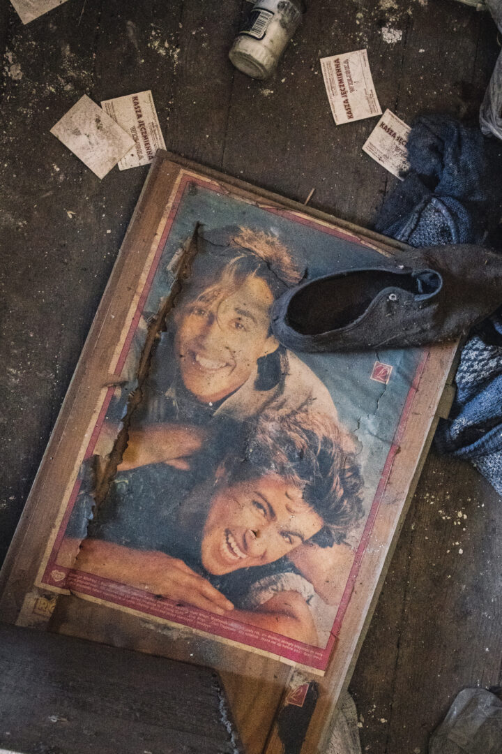 A torn poster of a smiling couple lying on a dusty floor, with a black boot resting on the frame and scattered papers nearby.