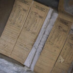 Old yellowed accounting forms (OPLATA TARGOWA) with columns and roman numerals, lying on a dusty concrete surface.