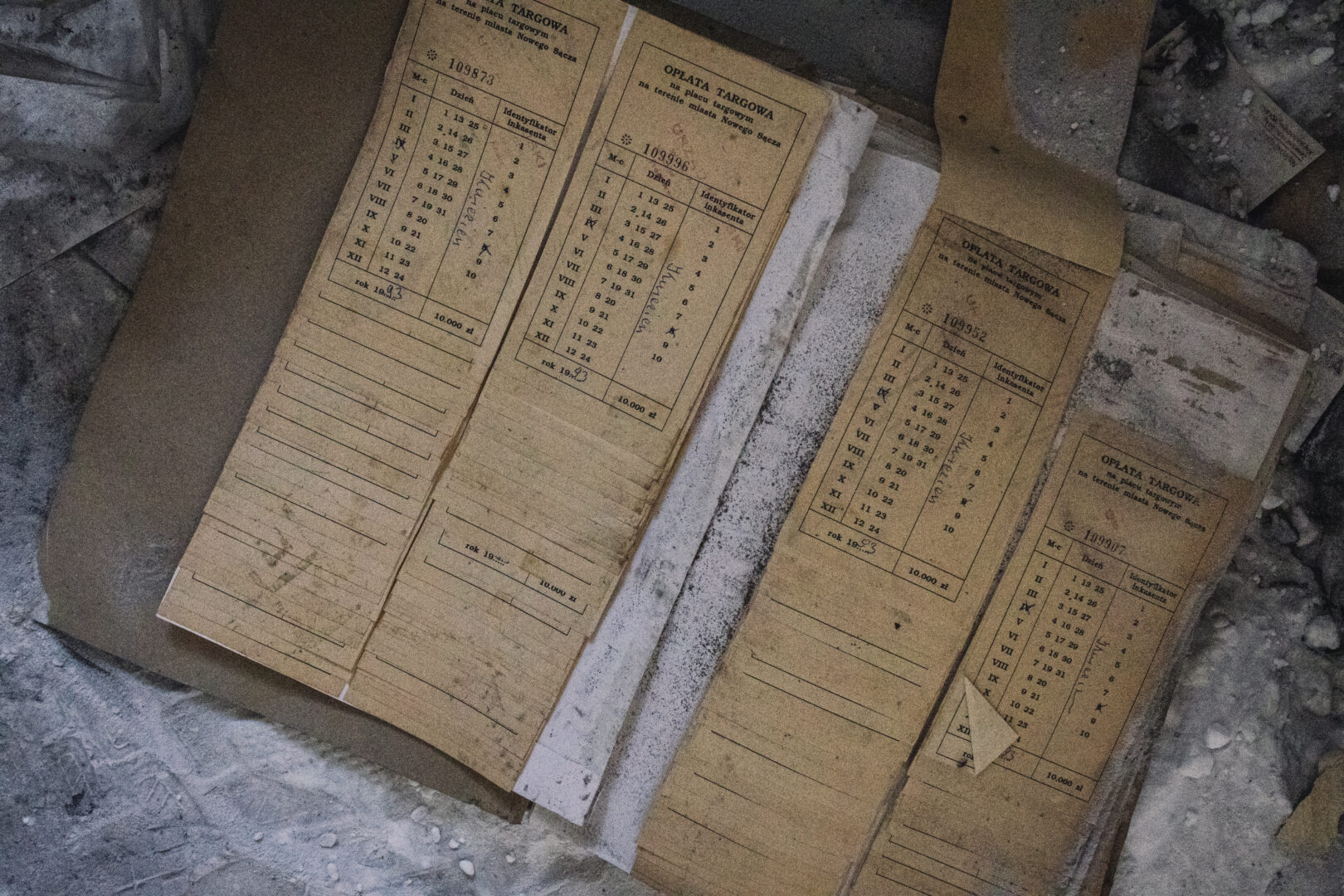 Old yellowed accounting forms (OPLATA TARGOWA) with columns and roman numerals, lying on a dusty concrete surface.