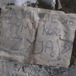 Dusty floor with a large cardboard sheet covered in black graffiti amid rubble and dirt in an abandoned space.