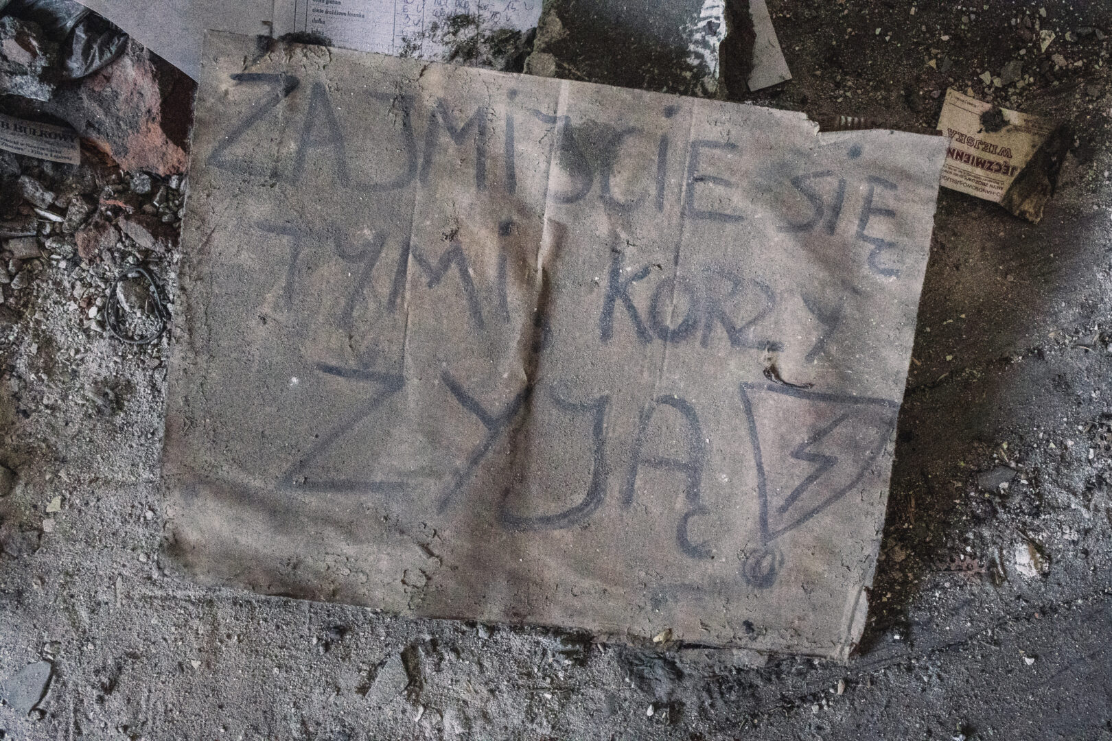 Dusty floor with a large cardboard sheet covered in black graffiti amid rubble and dirt in an abandoned space.