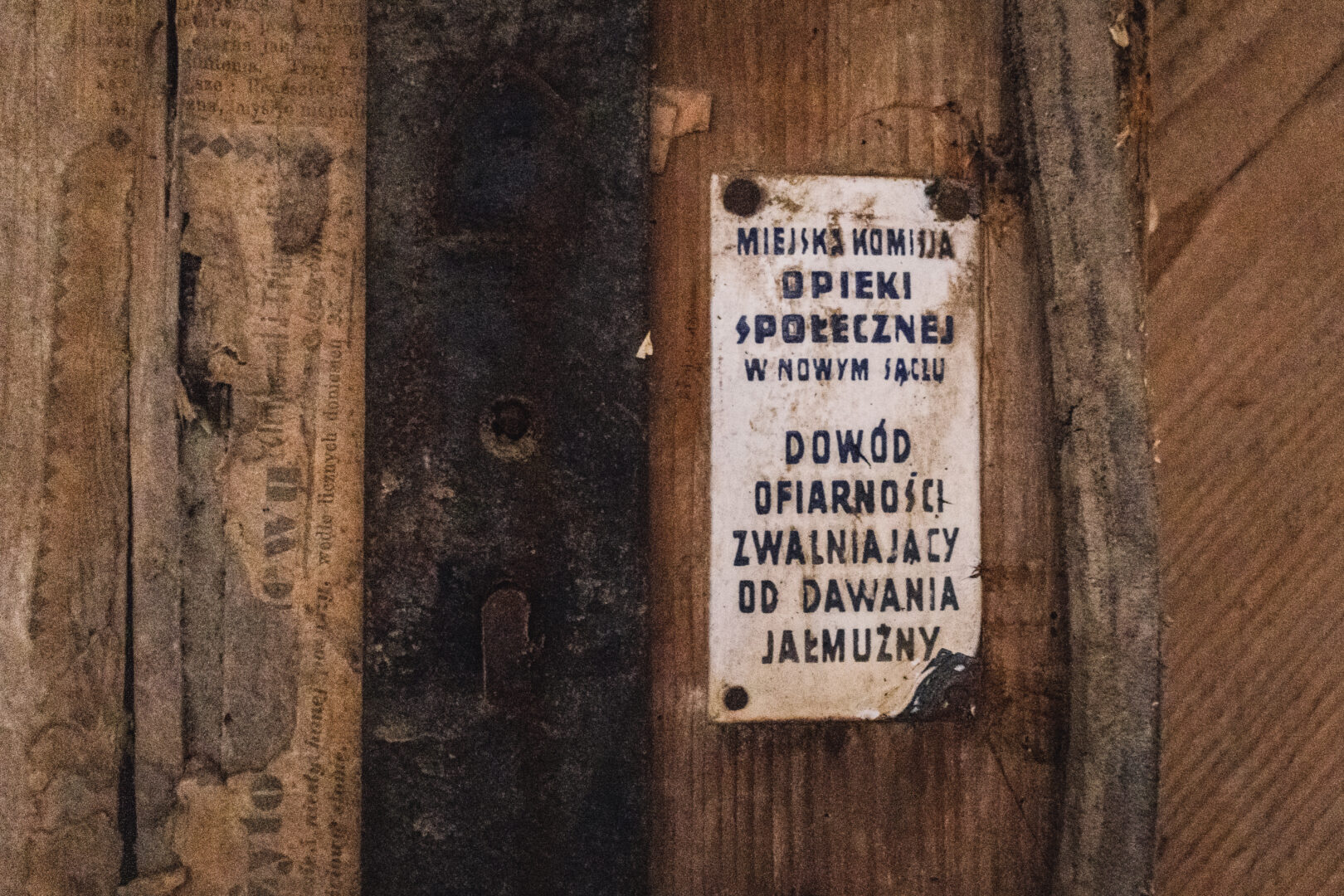 A weathered sign mounted on a wooden wall with Polish text about municipal social care and almsgiving.