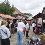 Crowd at an outdoor market along a cobblestone street, browsing items under wooden stalls with reddish roofs.