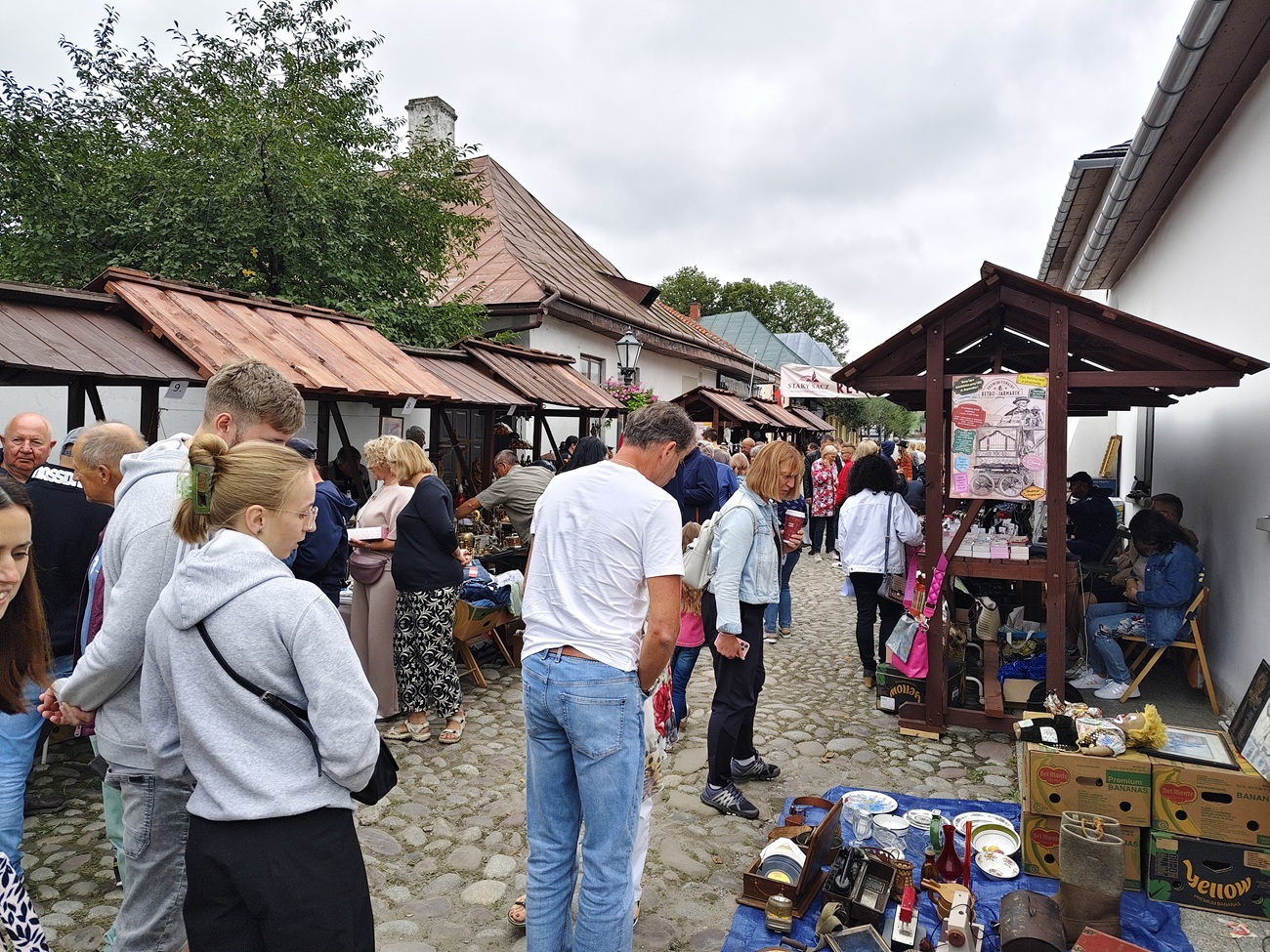 Crowd at an outdoor market along a cobblestone street, browsing items under wooden stalls with reddish roofs.