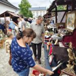 Woman in a blue patterned shirt inspecting vintage items laid out on a market table at an outdoor flea market on a cobblestone street.