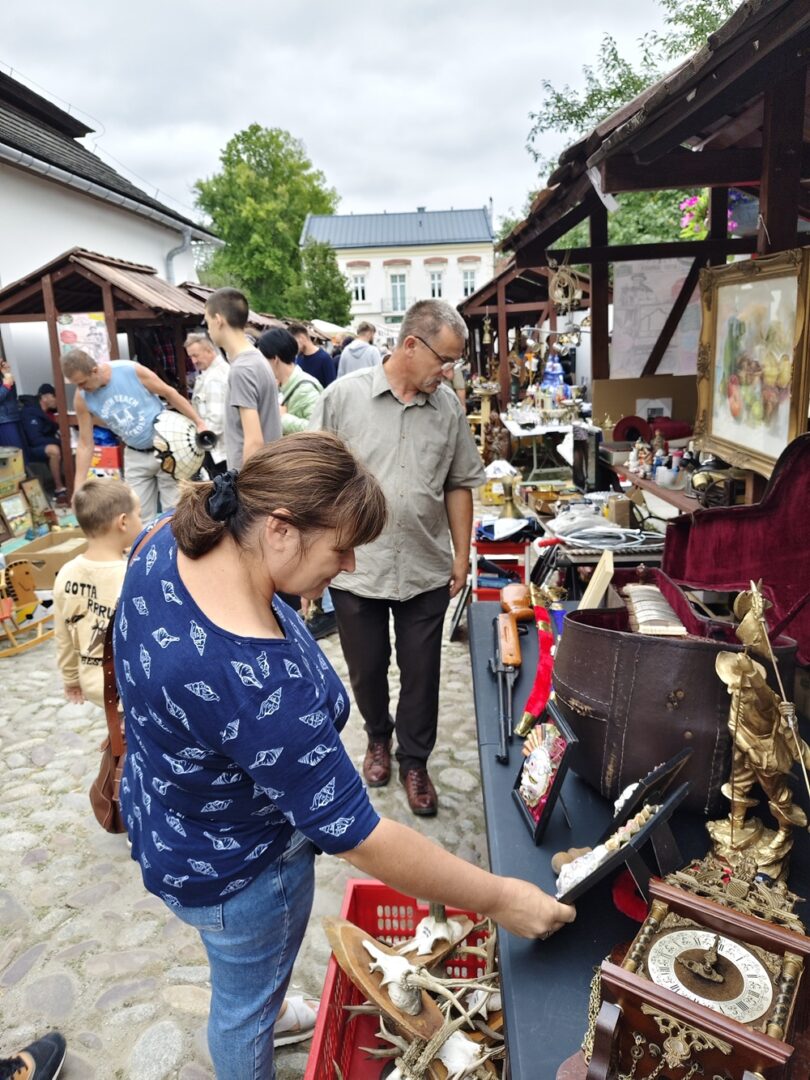 Woman in a blue patterned shirt inspecting vintage items laid out on a market table at an outdoor flea market on a cobblestone street.