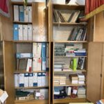 Wooden cabinet filled with blue and white binders and stacks of books in a cluttered storage room.