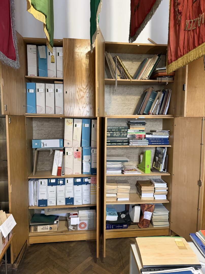 Wooden cabinet filled with blue and white binders and stacks of books in a cluttered storage room.