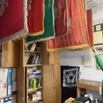 Row of ceremonial banners in red and green hanging from a rod over a desk, with shelves of books beneath.