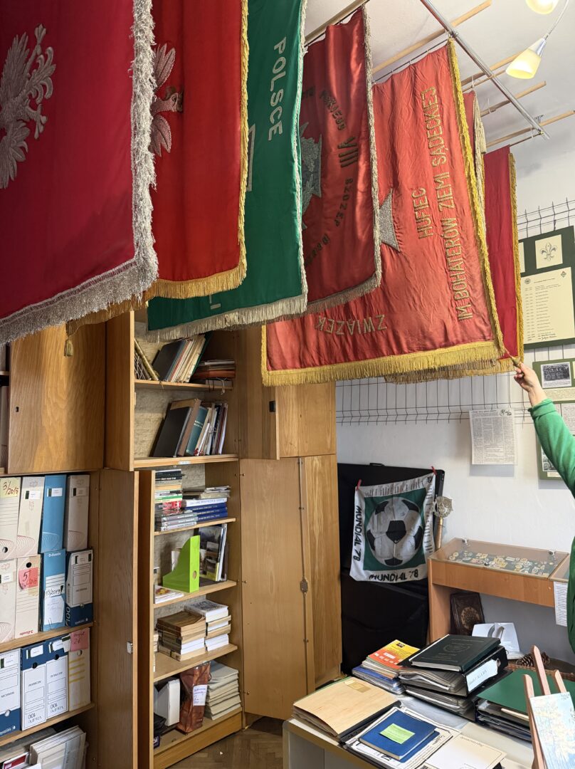 Row of ceremonial banners in red and green hanging from a rod over a desk, with shelves of books beneath.