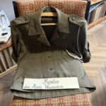 Olive-green military-style jacket draped over a chair with a handwritten sign displaying a name on its lapel area.