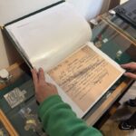 Person in a green sleeve opens a transparent display case to view an old handwritten document in a binder.