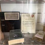 Display cabinet with awards and memorabilia: a framed certificate labeled Jubileusz Stulecia, a large black binder, and a dark green book titled Apel Podhalanski, plus a ceremonial banner on the right side.