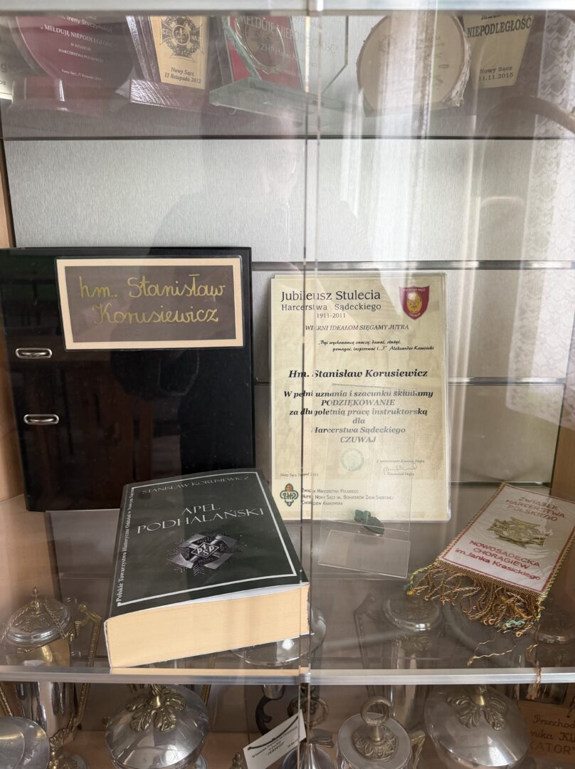 Display cabinet with awards and memorabilia: a framed certificate labeled Jubileusz Stulecia, a large black binder, and a dark green book titled Apel Podhalanski, plus a ceremonial banner on the right side.