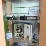 Glass display cabinet with badges, coins, medals, a white peaked cap, a scrapbook, and photos on three shelves.