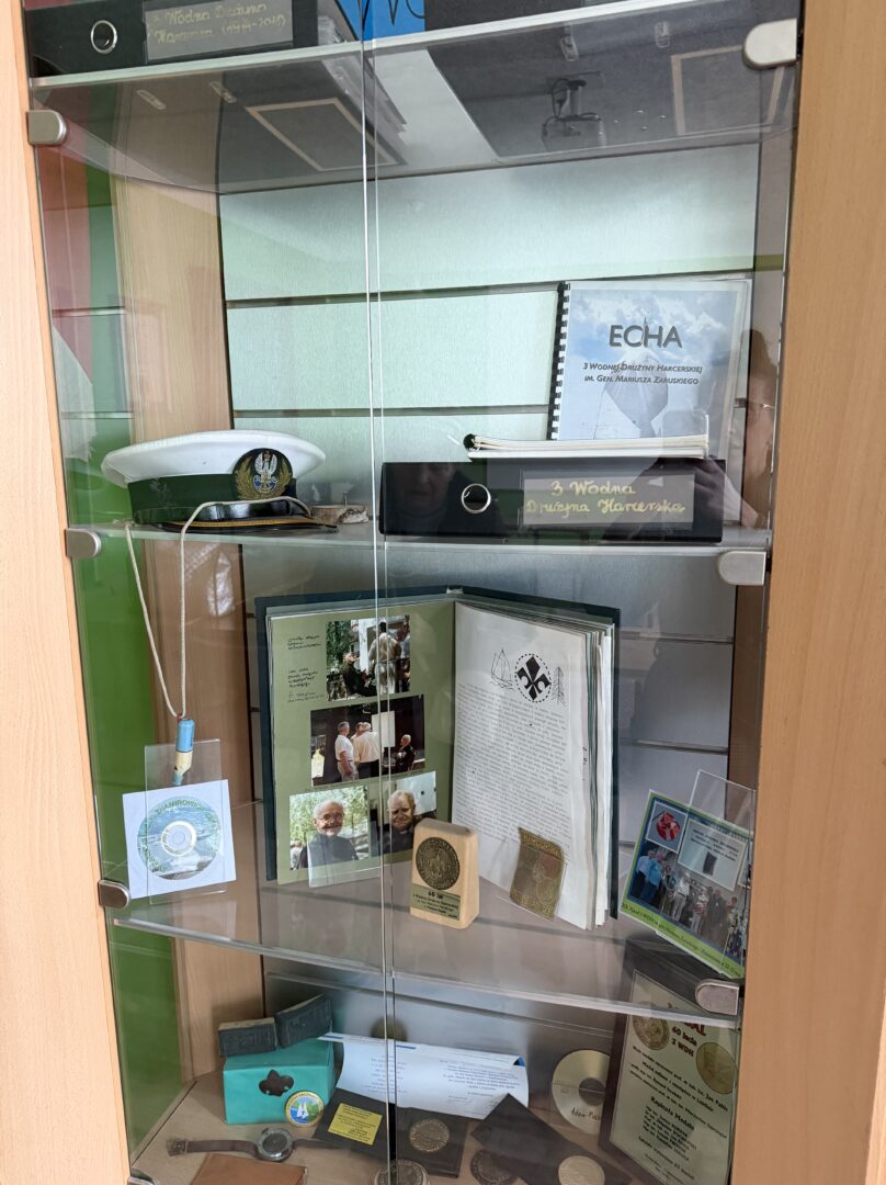 Glass display cabinet with badges, coins, medals, a white peaked cap, a scrapbook, and photos on three shelves.