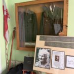 Glass-front wooden display case containing green military uniforms with medals and a hat, with a red-and-white flag beside it; a chalkboard-style exhibit board sits in front.