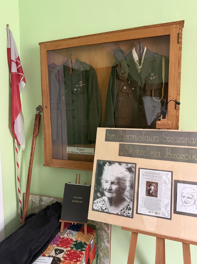 Glass-front wooden display case containing green military uniforms with medals and a hat, with a red-and-white flag beside it; a chalkboard-style exhibit board sits in front.