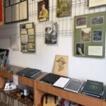 Wall displays with historical photos, plaques, and green panels in a museum case, above wooden shelves of books and artifacts.