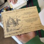 Beige vintage card with a detailed drawing of a wooden house and ornate border, held by a hand in a green sleeve. The text appears in decorative, Polish-style script at the top and along the page edge.