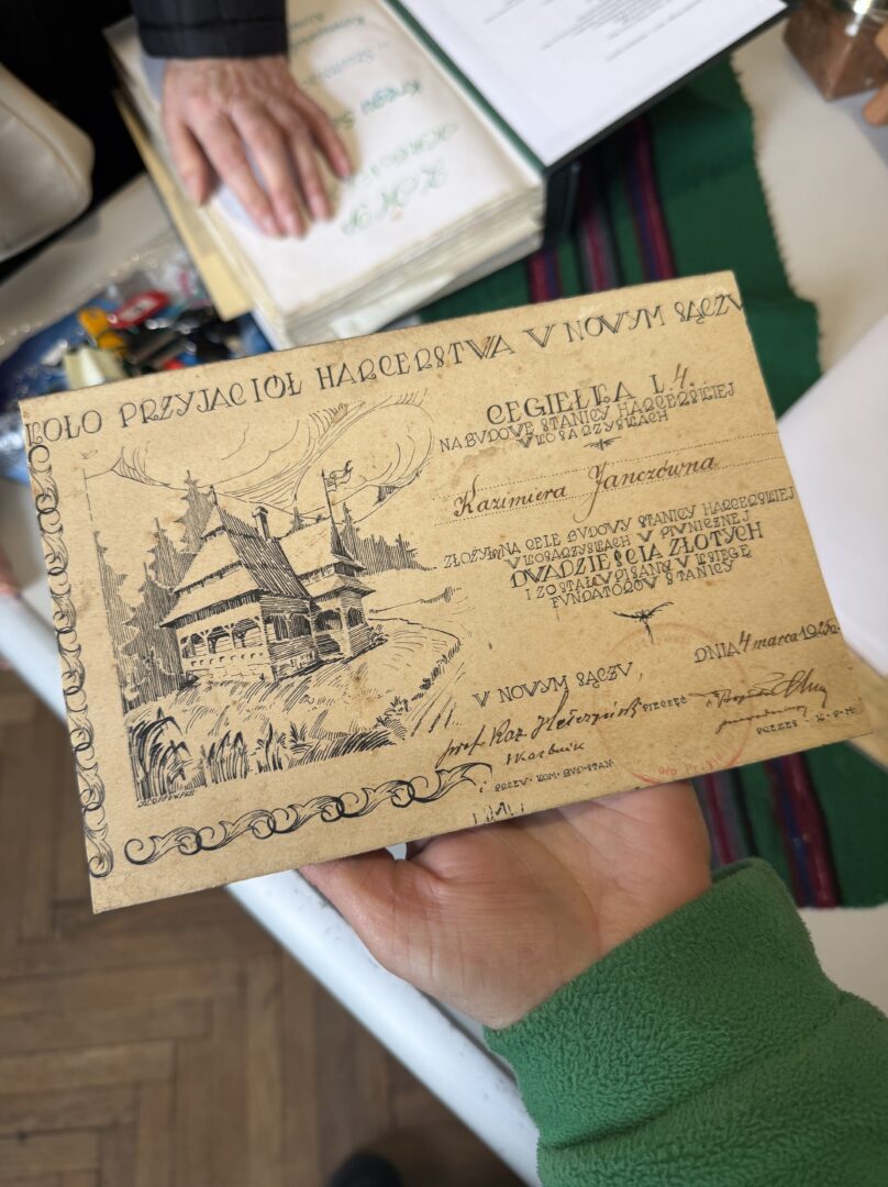 Beige vintage card with a detailed drawing of a wooden house and ornate border, held by a hand in a green sleeve. The text appears in decorative, Polish-style script at the top and along the page edge.