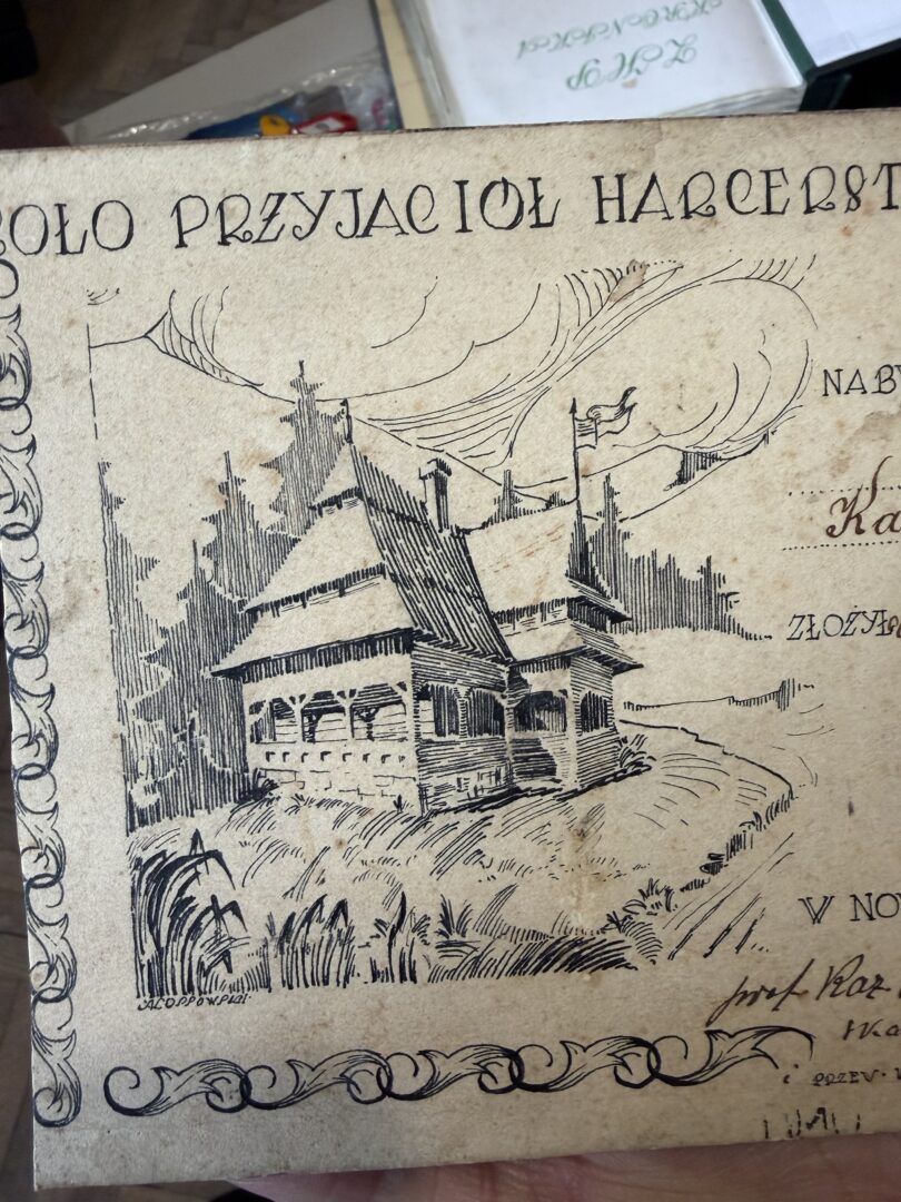 Vintage illustration of a quaint wooden cottage with a steep roof and wraparound porch, set among tall trees; decorative border and Polish text frame the scene.
