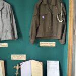 Olive green military-style jacket with patches on a hanger inside a green display case, with a white cord draped over the shoulder.
