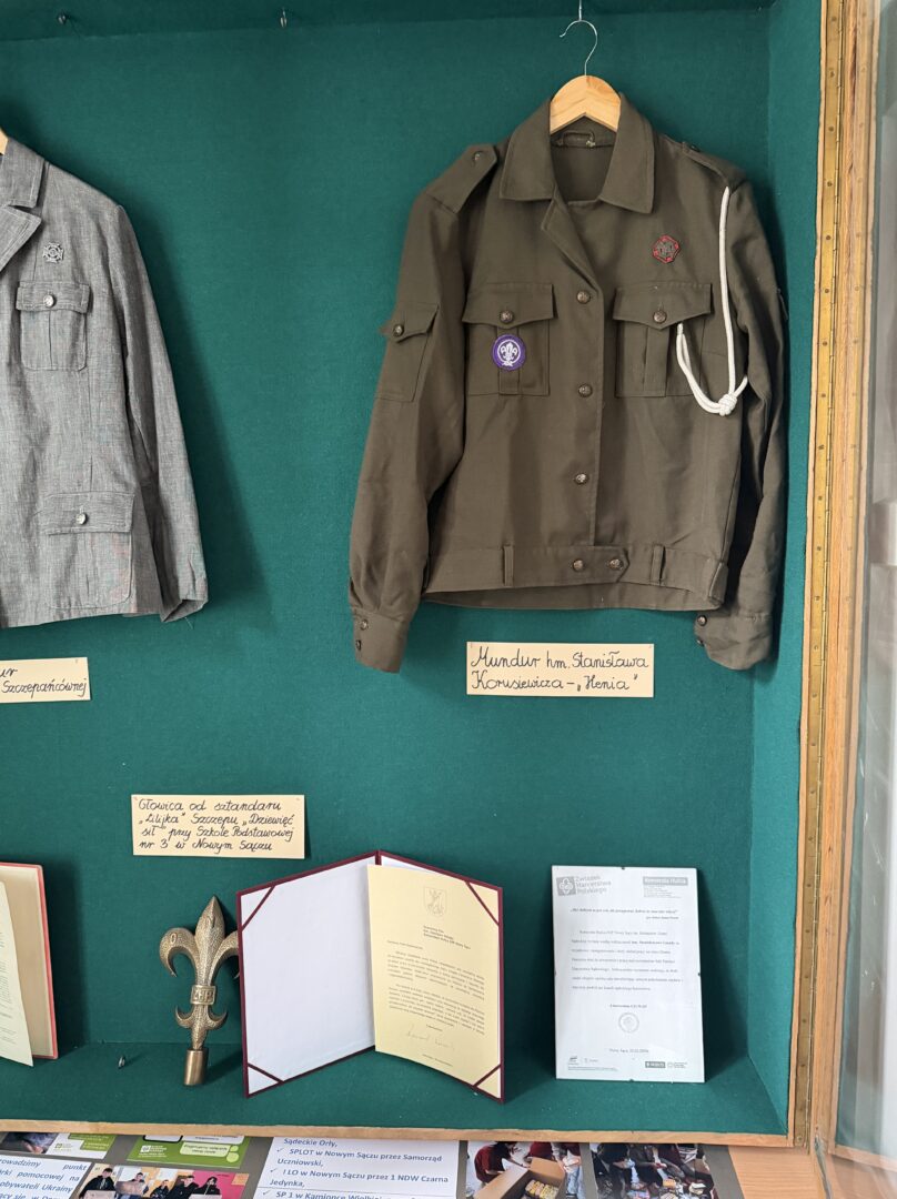 Olive green military-style jacket with patches on a hanger inside a green display case, with a white cord draped over the shoulder.