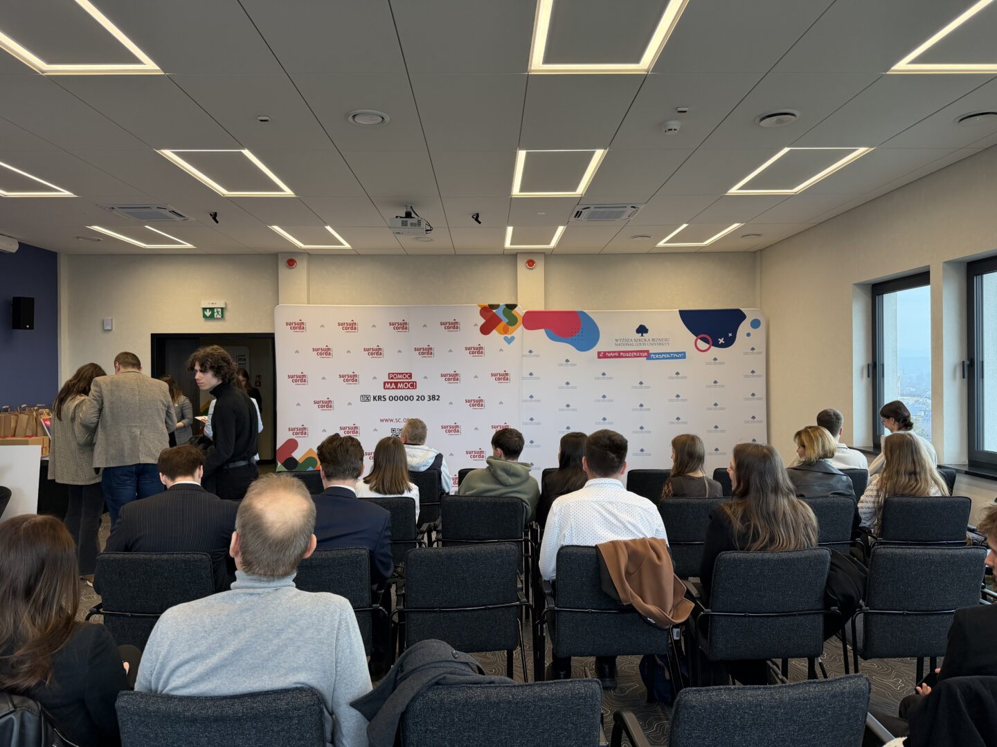 Audience seated in a modern conference room facing a sponsor backdrop with logos, windowed wall on the right, and people chatting at the left.