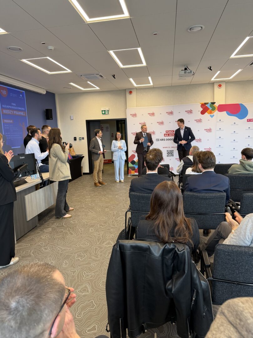 Four people on a stage presenting to an audience in a conference room, with a white logo backdrop behind them.