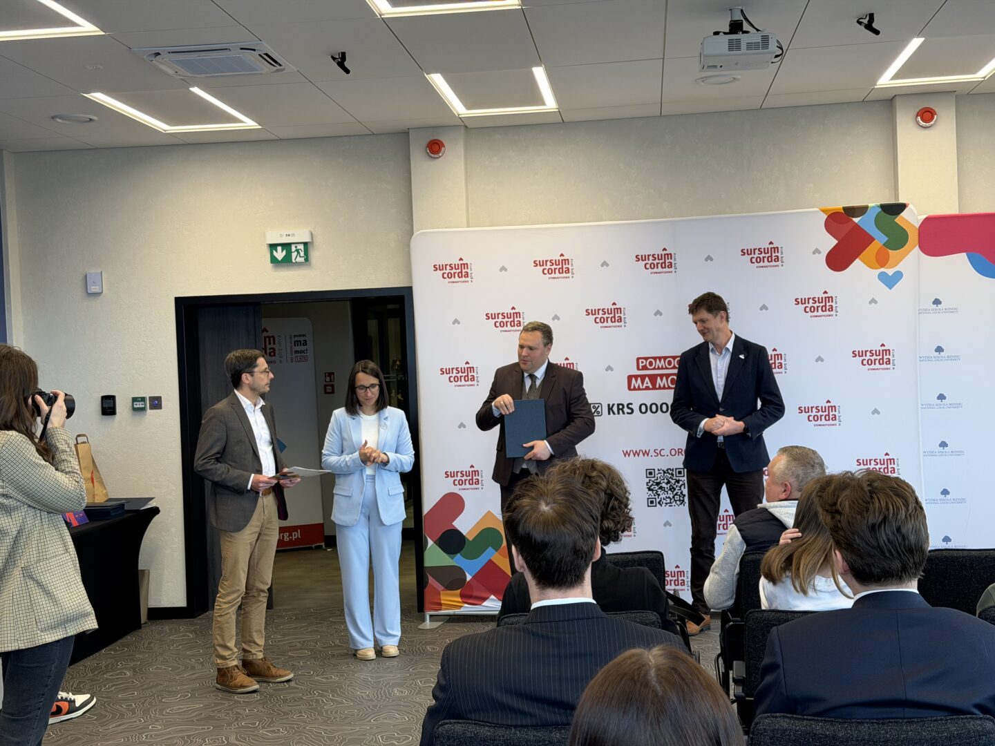 Panel of four people at a conference in front of a branded backdrop, one man presenting with a blue folder nearby other speakers in business attire.