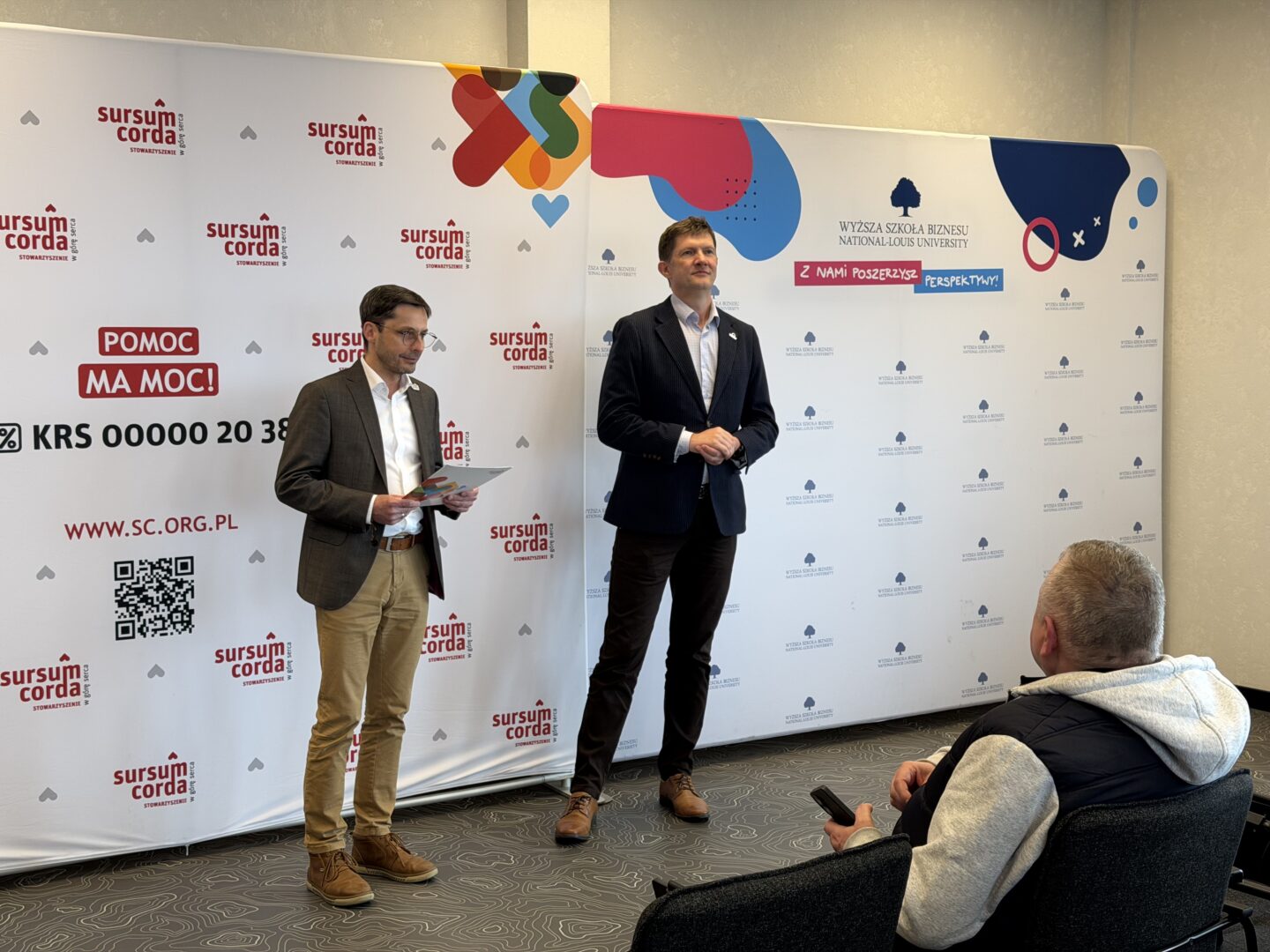 Two men in suits stand at a branded backdrop during a press event; one holds papers while the other speaks in front of logos.