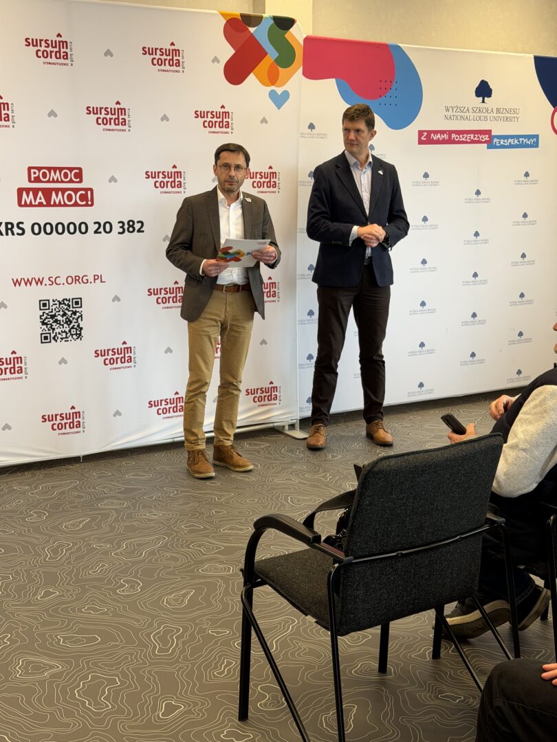 Two men in suits stand at a presentation backdrop with logos, one holding a folder of notes.
