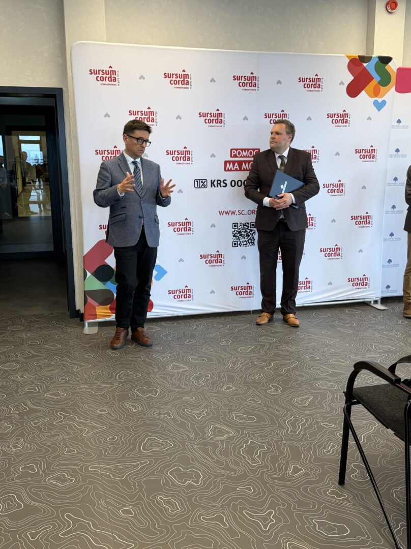 Two men in business attire stand in front of a branded backdrop at an event, one talking with hand gestures while the other holds a folder.