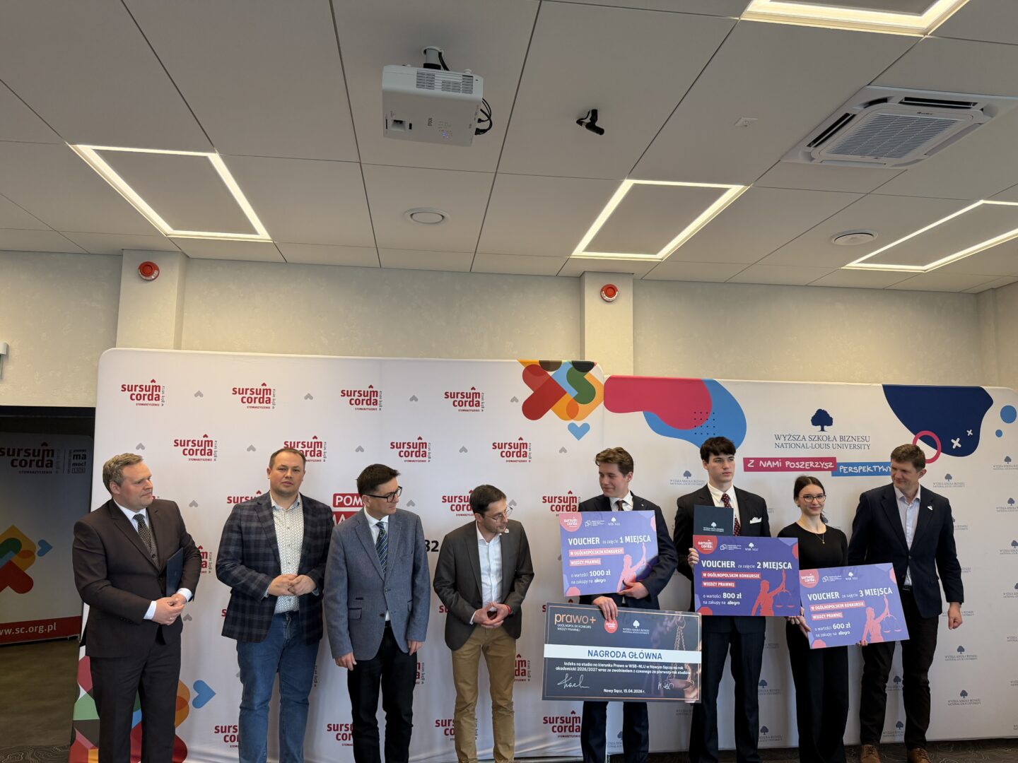 Group of seven people in business attire posing on stage with oversized vouchers and a large check, standing in front of a branded backdrop at an awards event.