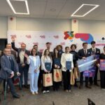 Group of young professionals posing for a ceremony photo, holding folders, gift bags, and oversized checks against a sponsor-branded backdrop.
