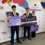 Three people in suits pose with oversized prize vouchers during an award ceremony.