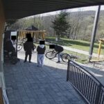 Group of people with bicycles standing on a paved patio outside a wooden building, with trees in the background.