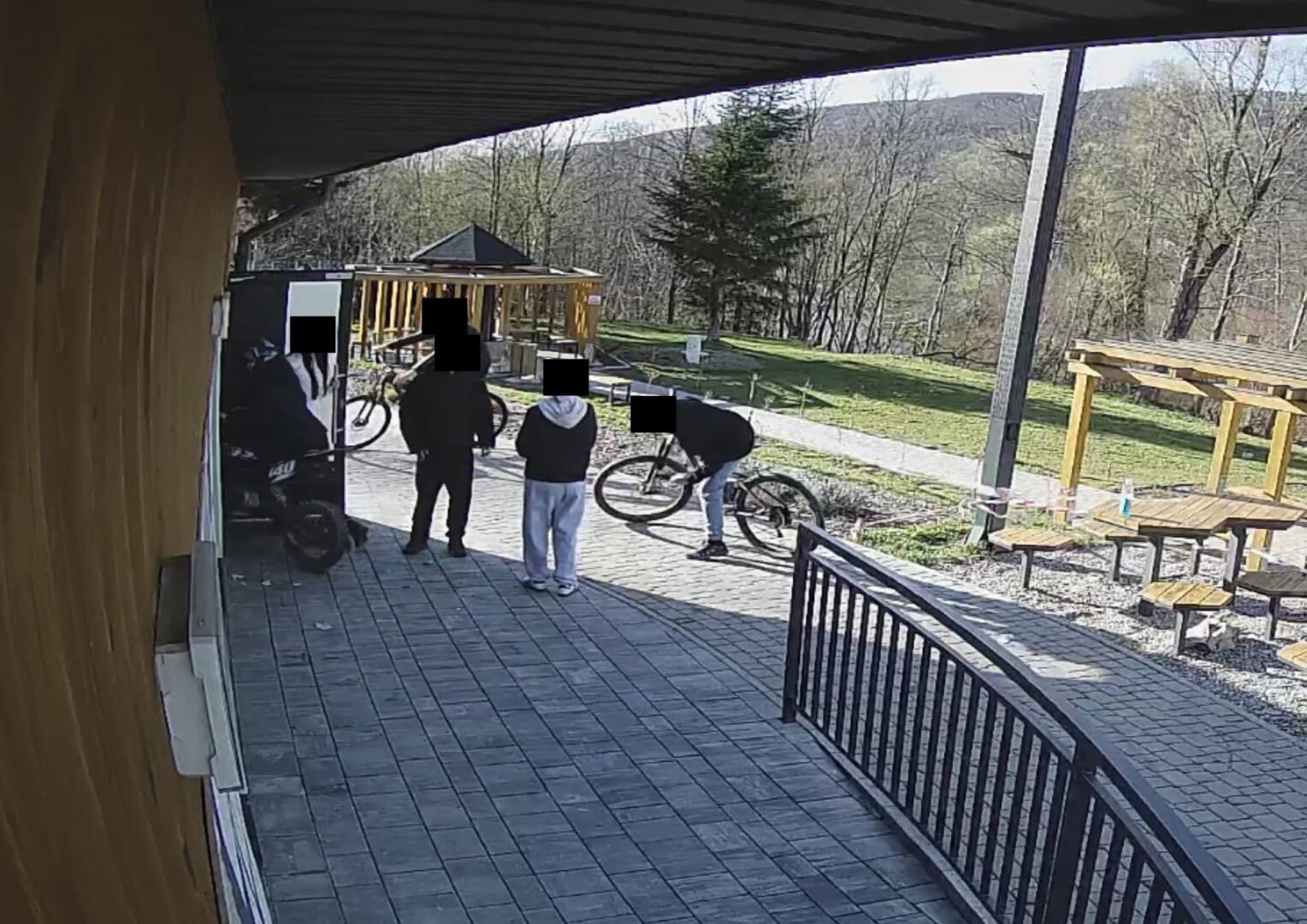 Group of people with bicycles standing on a paved patio outside a wooden building, with trees in the background.