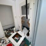 Interior doorway in a damaged room with a large hole in the wall, broken drywall, and scattered debris including a red bucket and a detached window frame on the floor.
