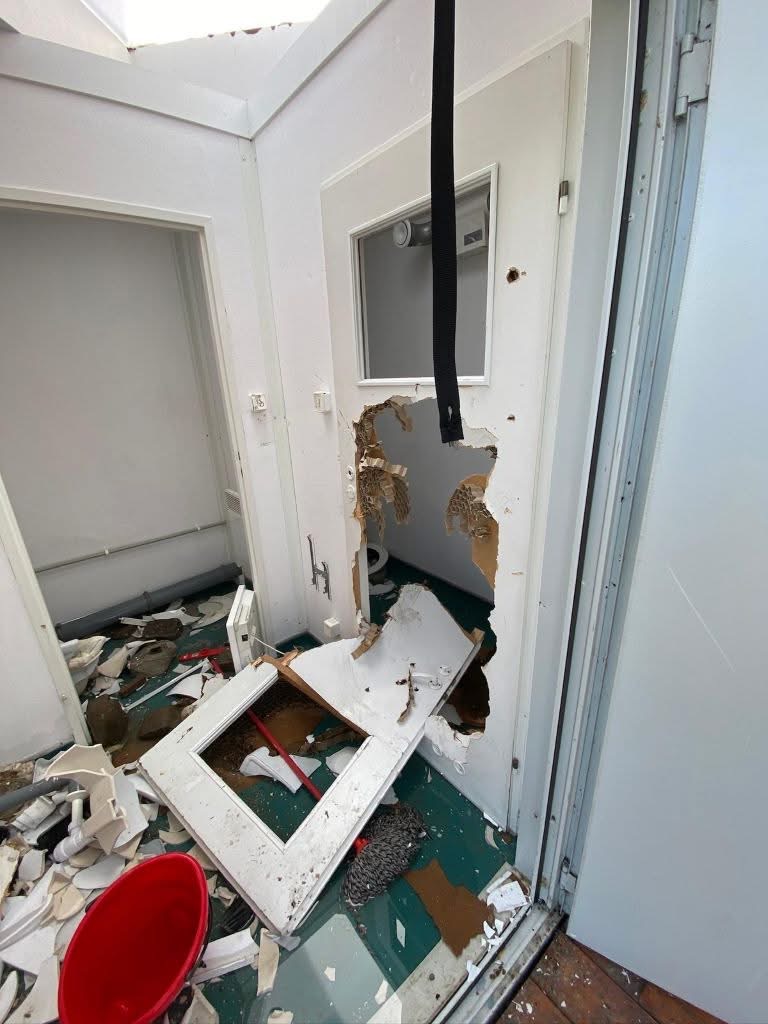 Interior doorway in a damaged room with a large hole in the wall, broken drywall, and scattered debris including a red bucket and a detached window frame on the floor.
