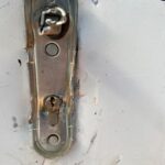 Close-up of a weathered door lock plate with a broken latch and keyhole on a gray-painted door frame.