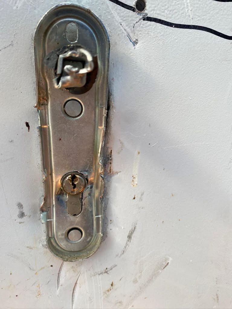 Close-up of a weathered door lock plate with a broken latch and keyhole on a gray-painted door frame.