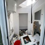 Interior of a severely damaged utility room: exposed pipes and a water heater on the left, torn drywall on the right, and debris scattered across a green floor with a red bucket in the mix.