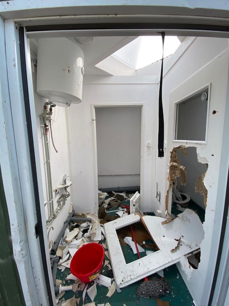Interior of a severely damaged utility room: exposed pipes and a water heater on the left, torn drywall on the right, and debris scattered across a green floor with a red bucket in the mix.