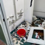 Messy demolition scene in a bathroom with exposed pipes, broken plaster, and a red bucket among debris.
