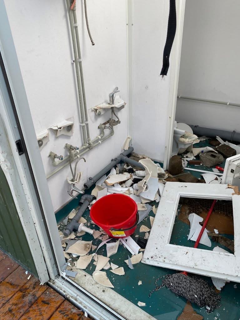 Messy demolition scene in a bathroom with exposed pipes, broken plaster, and a red bucket among debris.